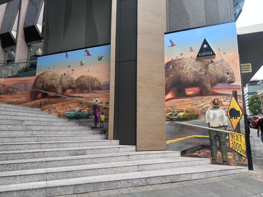 This image shows a large-scale public mural displayed on the exterior walls of a building. The mural depicts a surreal, desert-like landscape with three large, furry wombats walking across the landscape. The wombats are surrounded by flying birds and cars. There are people standing in the foreground facing the wombats. There are stairs and ramps leading up to the building. Overall, the artwork has a fantastical, whimsical quality that draws the viewer's attention.